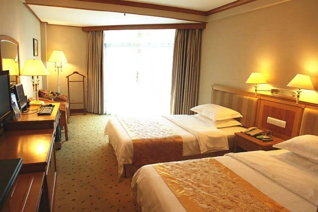 A hotel room with two beds, a desk with a computer, lamps, and a large window with curtains.