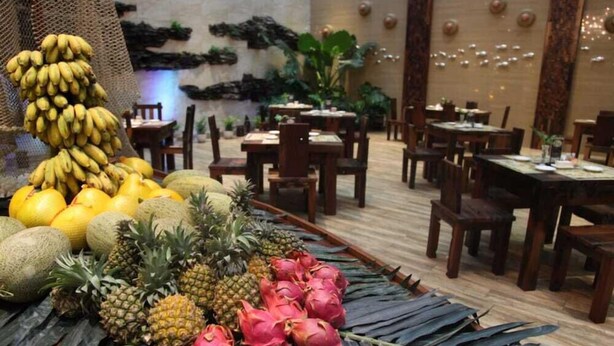 Un restaurante con mesas y sillas de madera, una exhibición de frutas con plátanos, limones y pitahaya, y una pared decorativa con plantas e iluminación.
