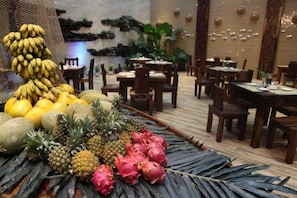 Un restaurante con mesas y sillas de madera, una exhibición de frutas con plátanos, limones y pitahaya, y una pared decorativa con plantas e iluminación.