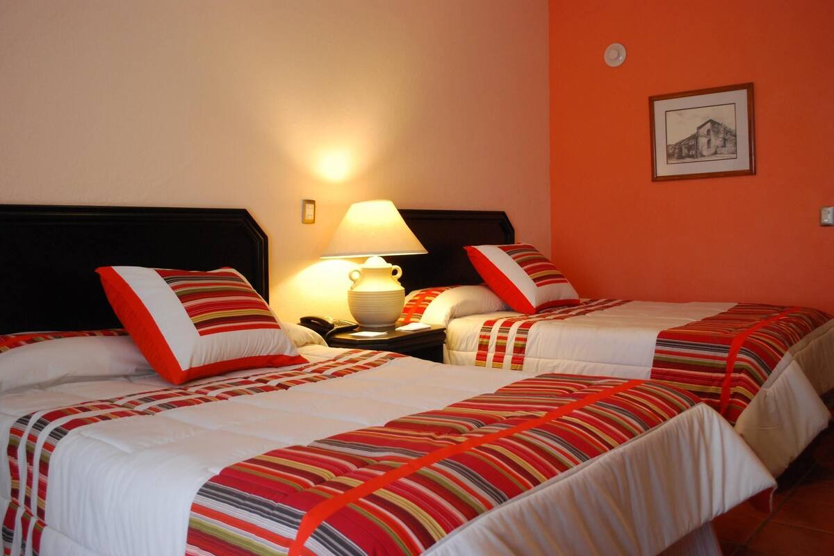 Ein Hotelzimmer mit zwei Betten, jeweils mit gestreifter Bettwäsche und roten Akzenten. An einem Nachttisch steht eine Lampe und ein Telefon.