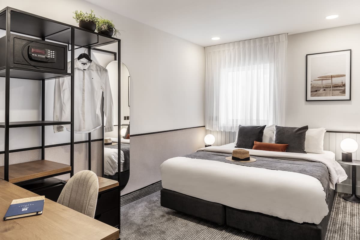 A hotel room with a bed, bedside tables, a desk, a chair, a wardrobe, and a window with curtains.