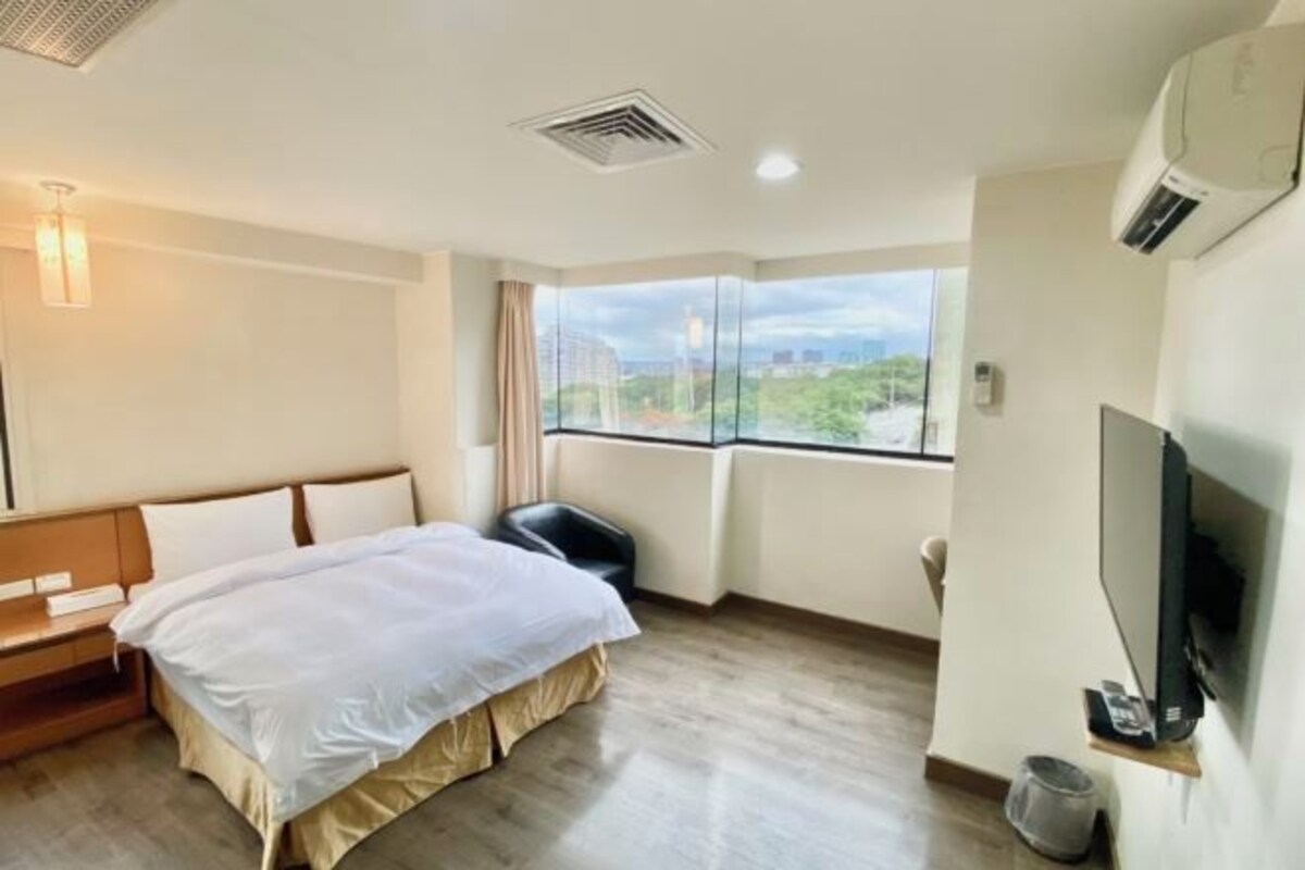 A hotel room with a bed, a TV, a window with a view, and a ceiling vent.