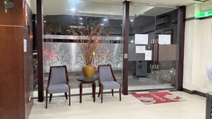 Uma área de recepção com porta de vidro, duas cadeiras de madeira, uma mesa pequena e um vaso com plantas secas.