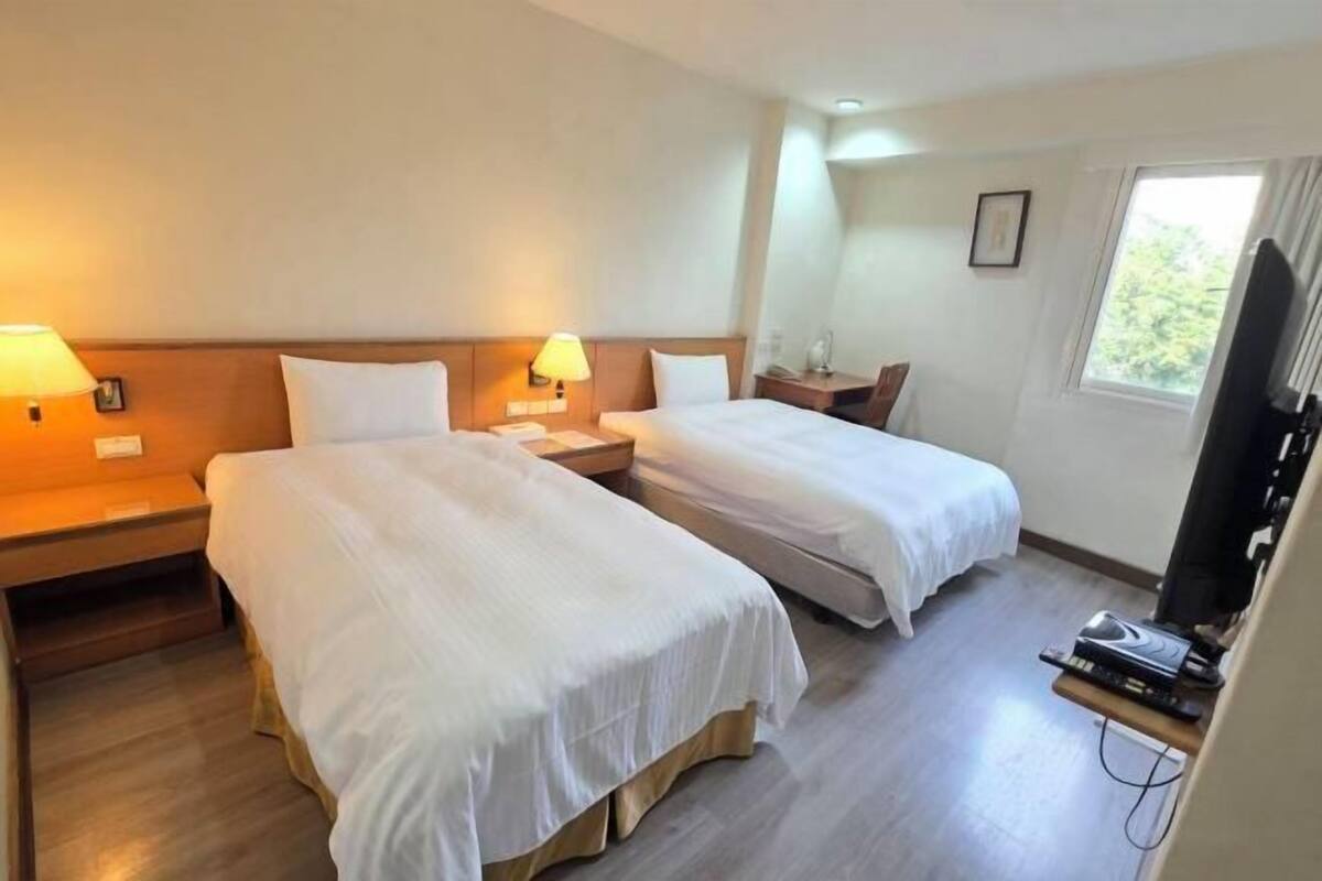 A hotel room with two beds, bedside tables, lamps, a television, and a window.