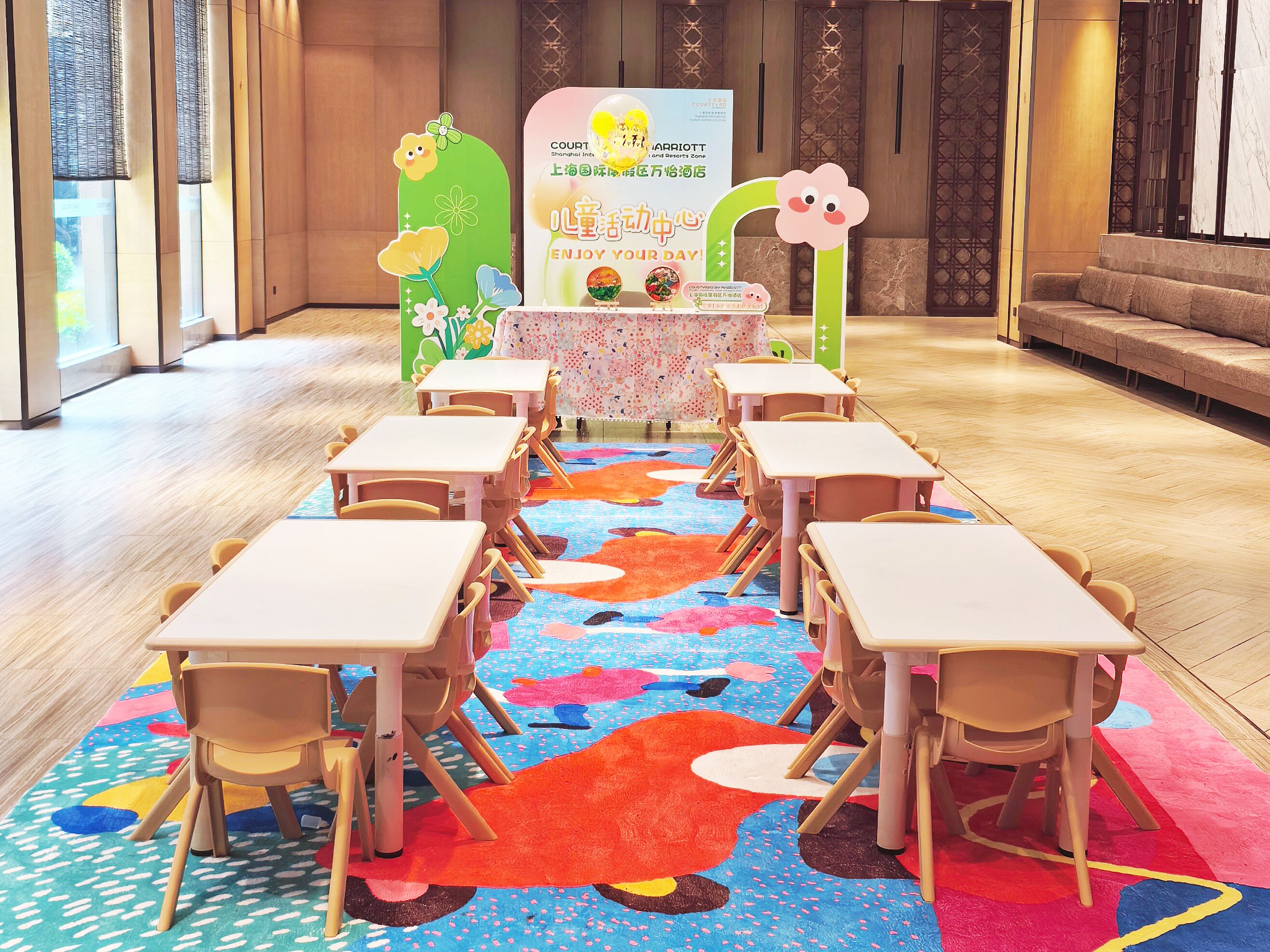 A children's play area with tables and chairs arranged in rows, a colorful carpet, and a decorative backdrop with a flower and a sign.