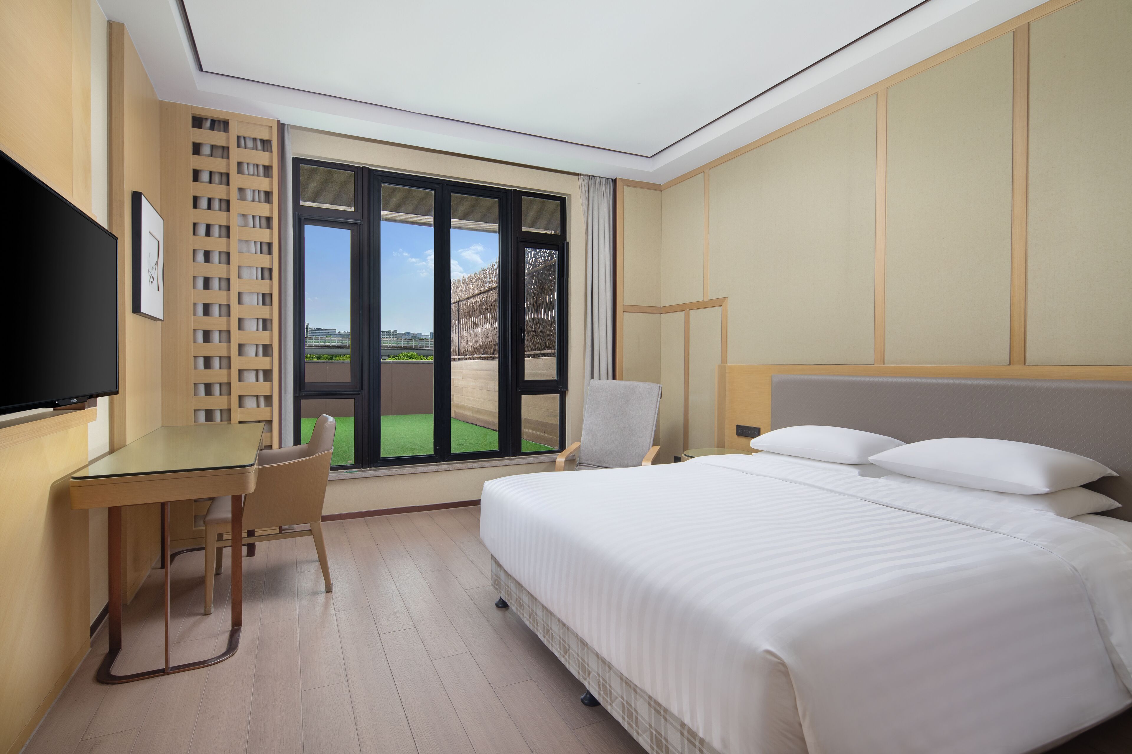 A hotel room with a bed, a desk, a chair, a TV, and a large window with a view.