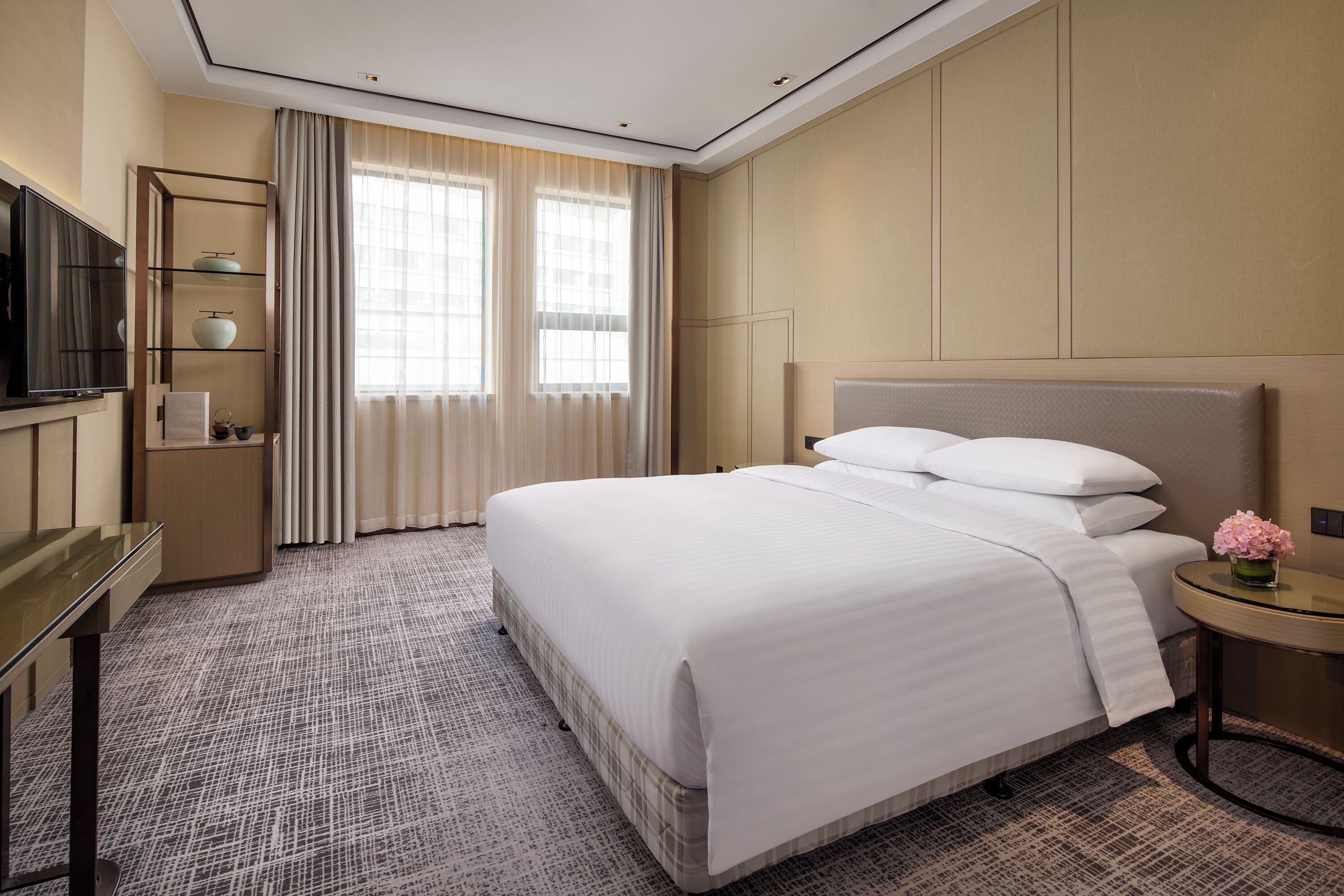 A hotel room with a large bed, a TV on a stand, a small table with a vase of flowers, and a window with curtains.