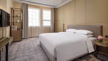 A hotel room with a large bed, a TV on a stand, a small table with a vase of flowers, and a window with curtains.