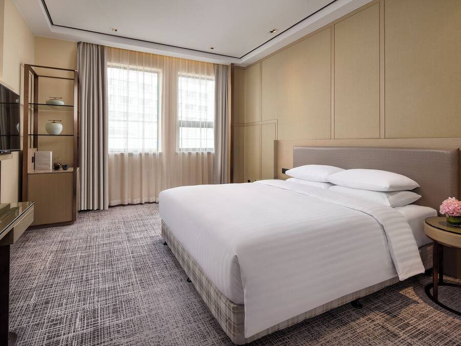 A hotel room with a large bed, a TV on a stand, a small table with a vase of flowers, and a window with curtains.