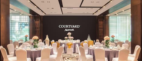 Un salón de conferencias con una pantalla grande que muestra 'COURTYARD Marriott' al frente. Las mesas están dispuestas con sillas blancas y centros de mesa.