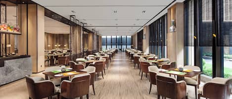 Un amplio salón comedor de hotel con largas mesas y sillas, una recepción y grandes ventanales que ofrecen vistas.