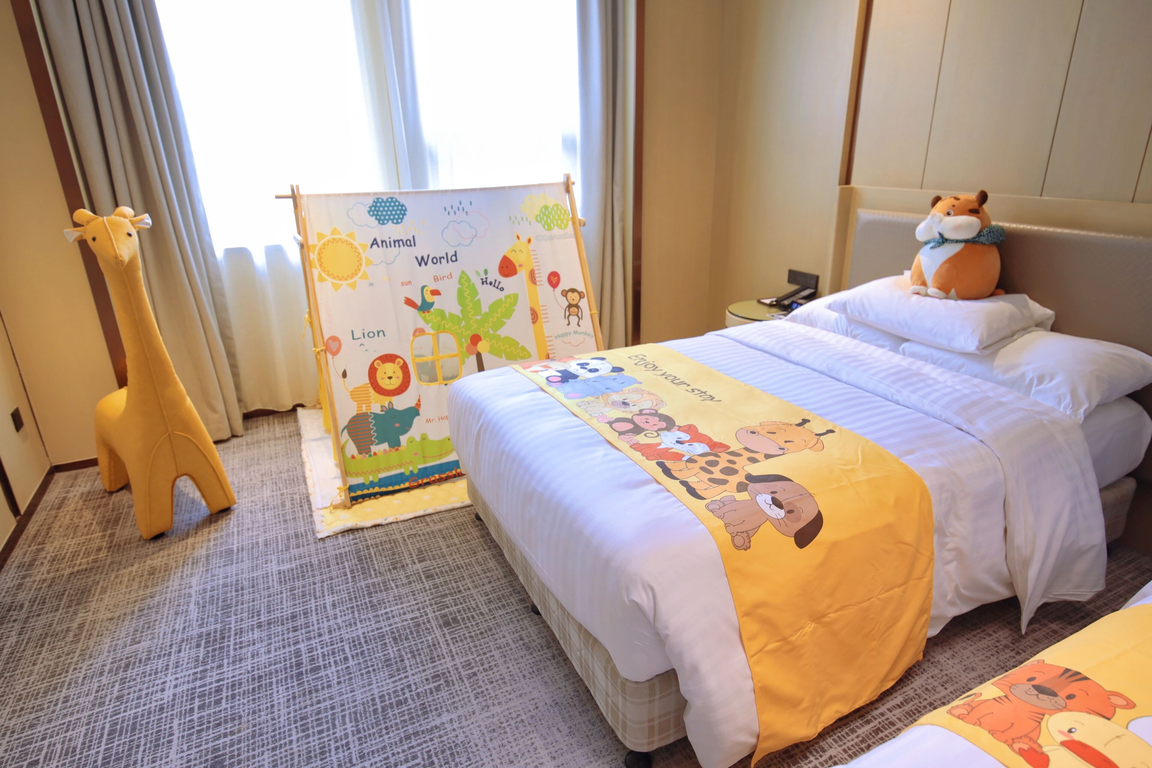 Een hotelkamer met een bed, een giraf knuffel, een educatief poster voor kinderen en een kussen met een tekenfilmfiguur.