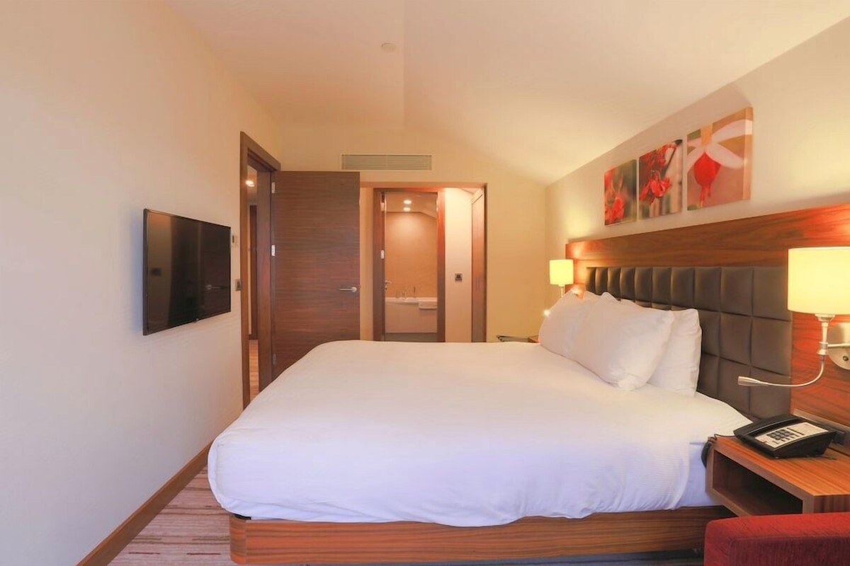 Una camera d'hotel con un letto grande, una televisione a parete, un comodino con una lampada e una porta che dà accesso al bagno.