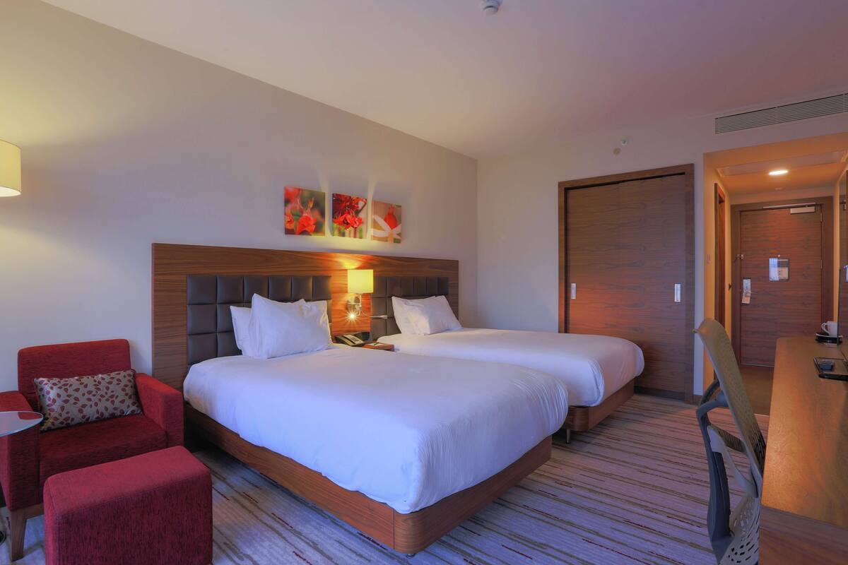 Habitación de hotel con dos camas, un sillón rojo, una mesita y una puerta de madera.