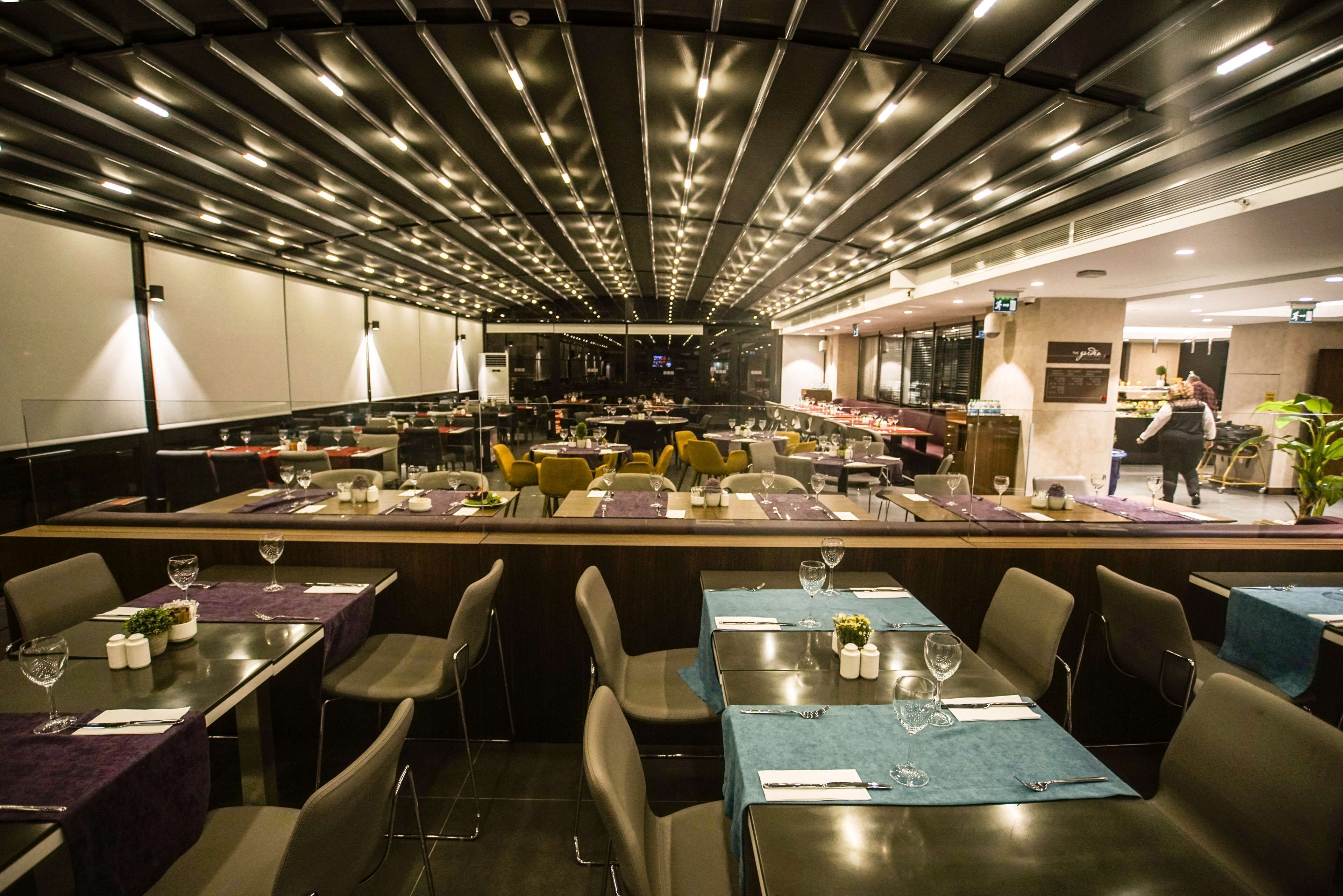 A modern restaurant with multiple dining tables, each set with cutlery and glassware, and a bar area with a bartender.