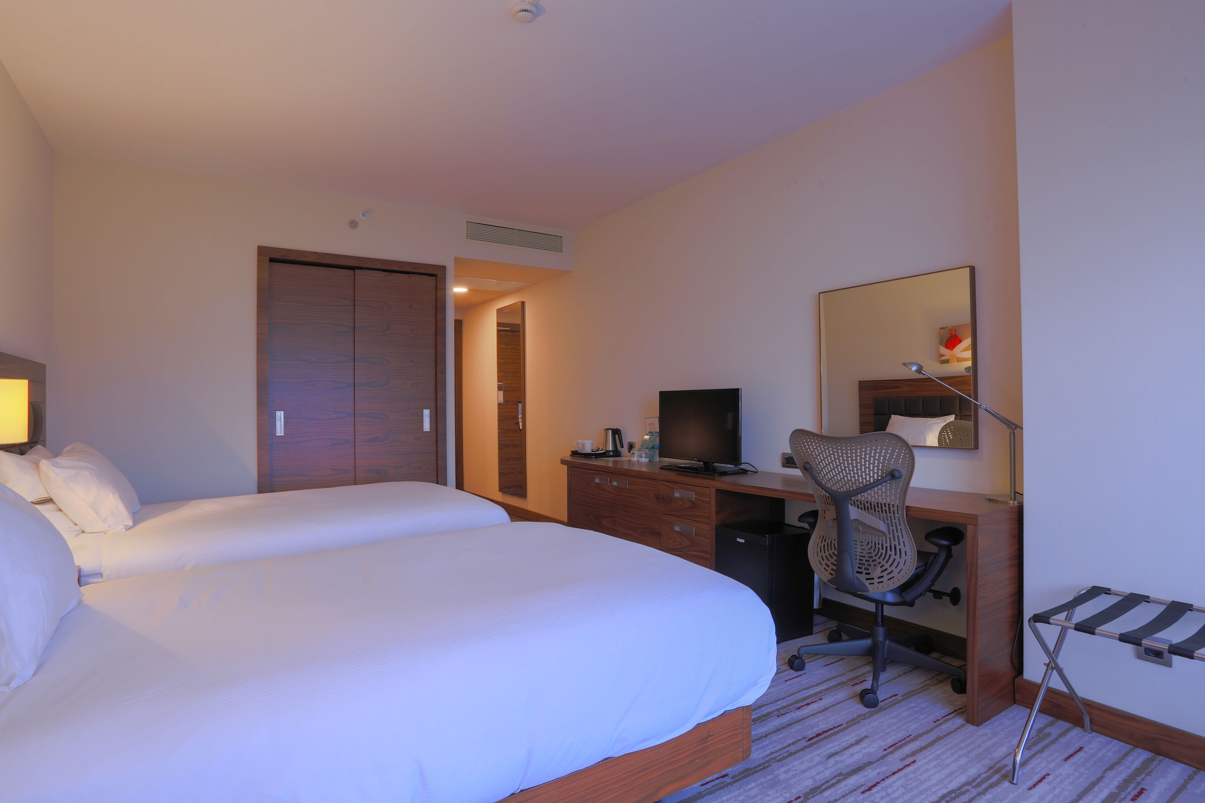 A hotel room with two beds, a desk with a computer, a chair, a TV, and a mirror.