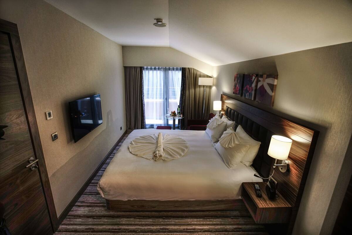 Habitación de hotel con una cama grande, un televisor de pantalla plana en la pared, una mesita de noche con lámpara y una ventana con cortinas.