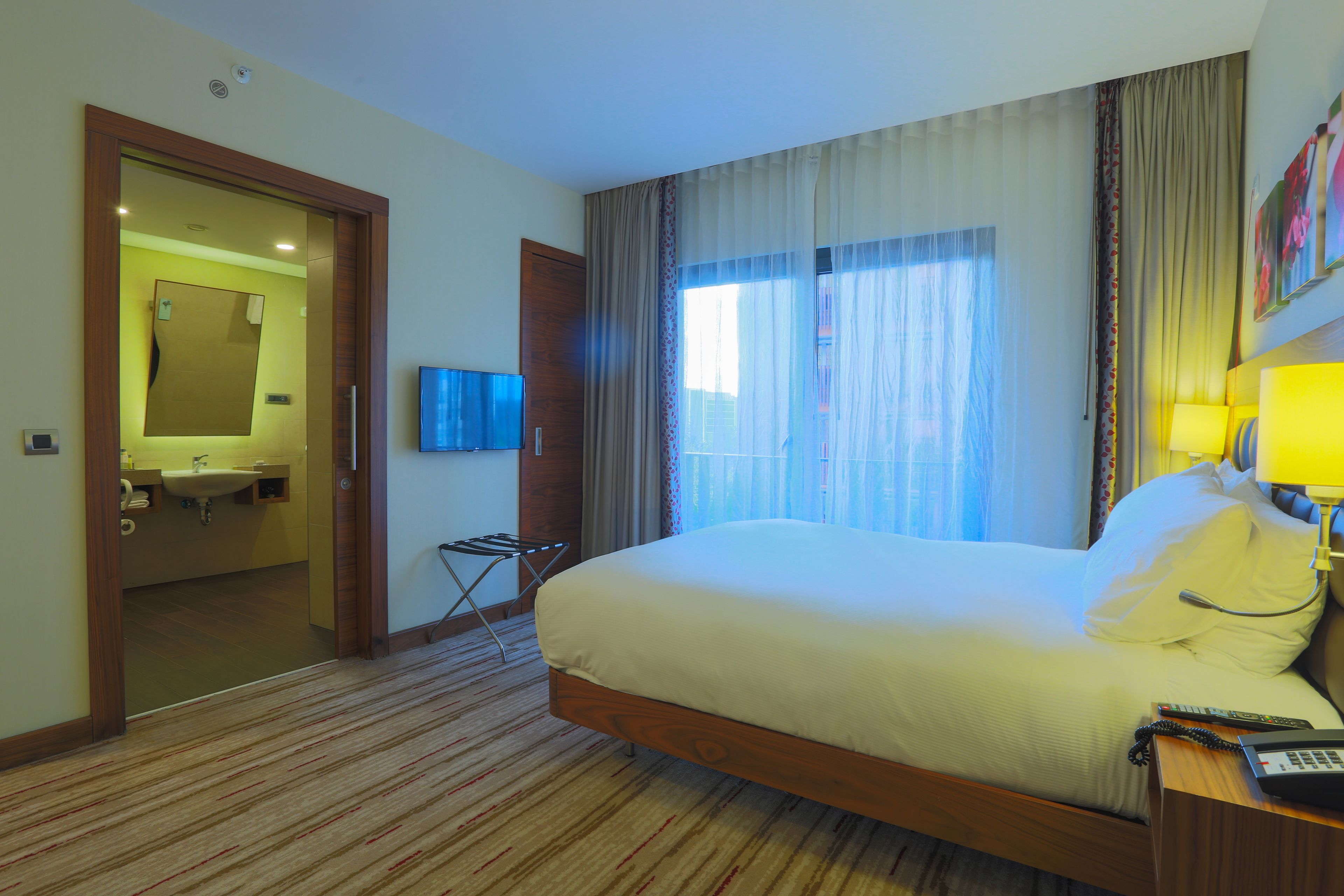 A hotel room with a large bed, a bedside table with a lamp, a TV, and a bathroom visible through an open door.