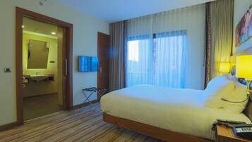 A hotel room with a large bed, a bedside table with a lamp, a TV, and a bathroom visible through an open door.