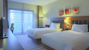 Habitación de hotel con dos camas, un escritorio con televisor, una lámpara y obras de arte en la pared.