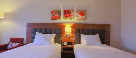 A hotel room with two beds, a nightstand with a lamp, and a red chair.