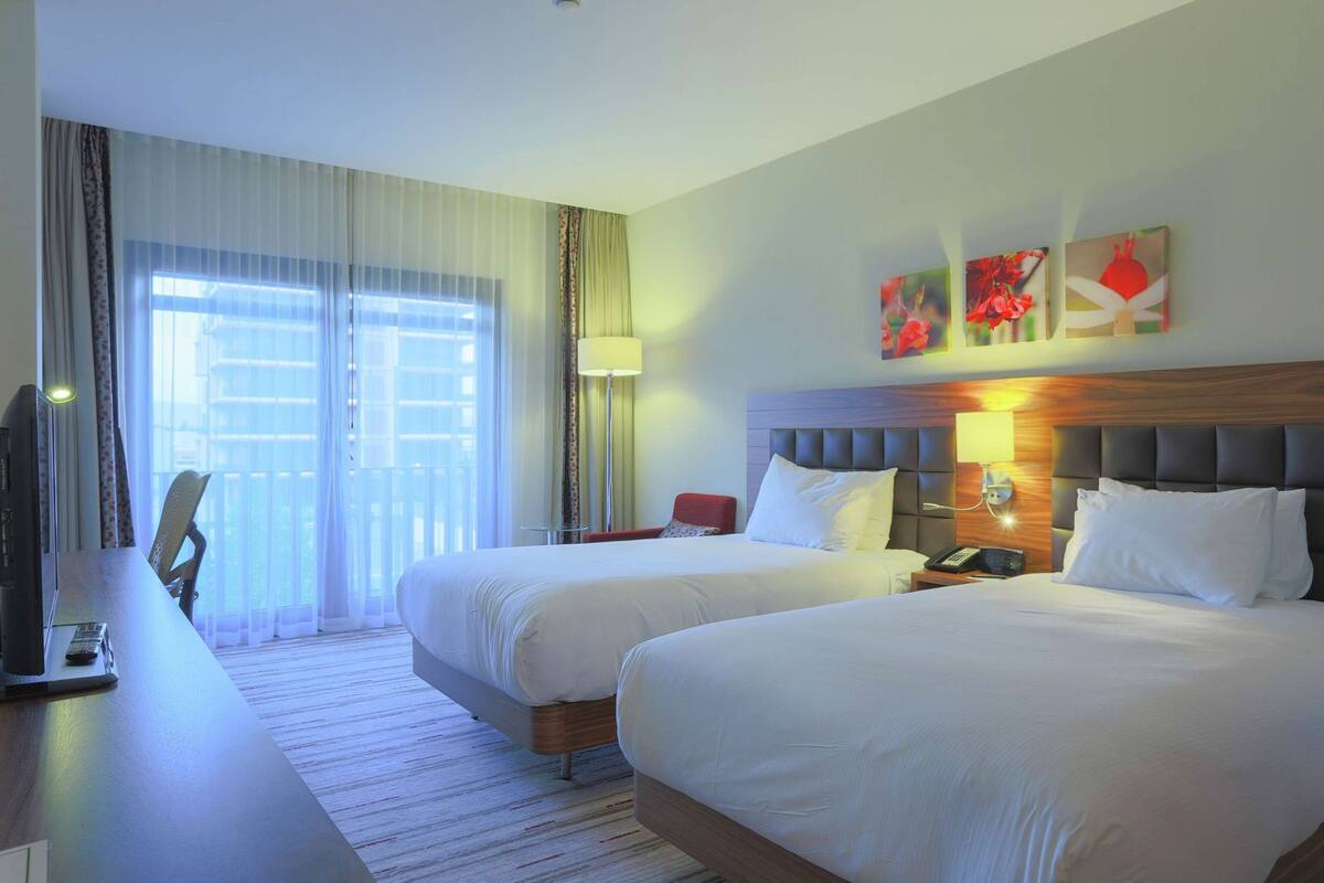 Habitación de hotel con dos camas, un escritorio con televisor, una silla y cuadros en la pared.