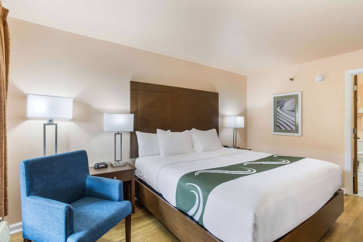 Ein Hotelzimmer mit einem großen Bett, zwei Nachttischlampen, einem blauen Sessel und einem gerahmten Bild an der Wand.