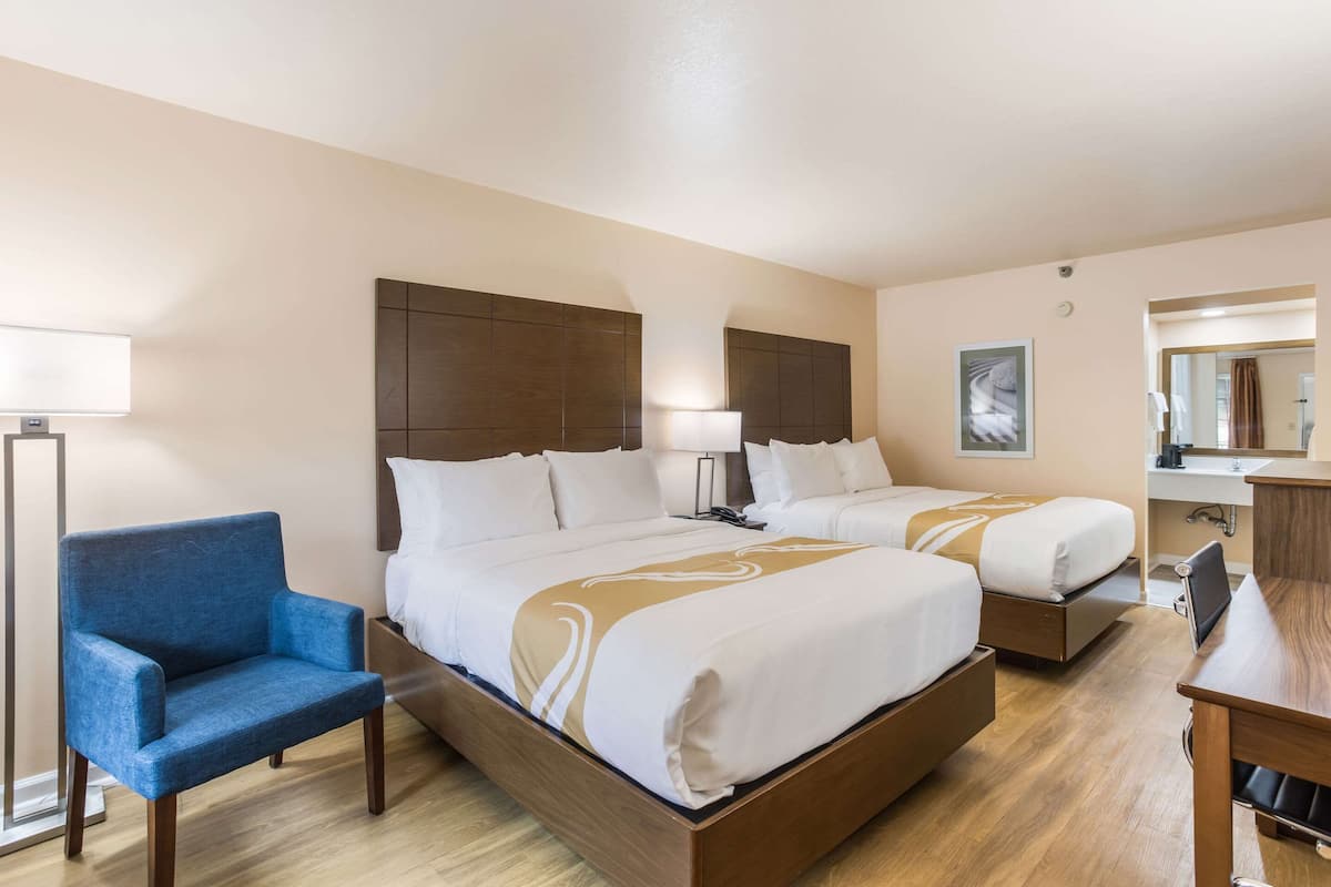 Ein Hotelzimmer mit zwei Betten, einem Schreibtisch, einem Stuhl, einer Lampe und einem durchgegangenen Tür sichtbaren Badezimmer.