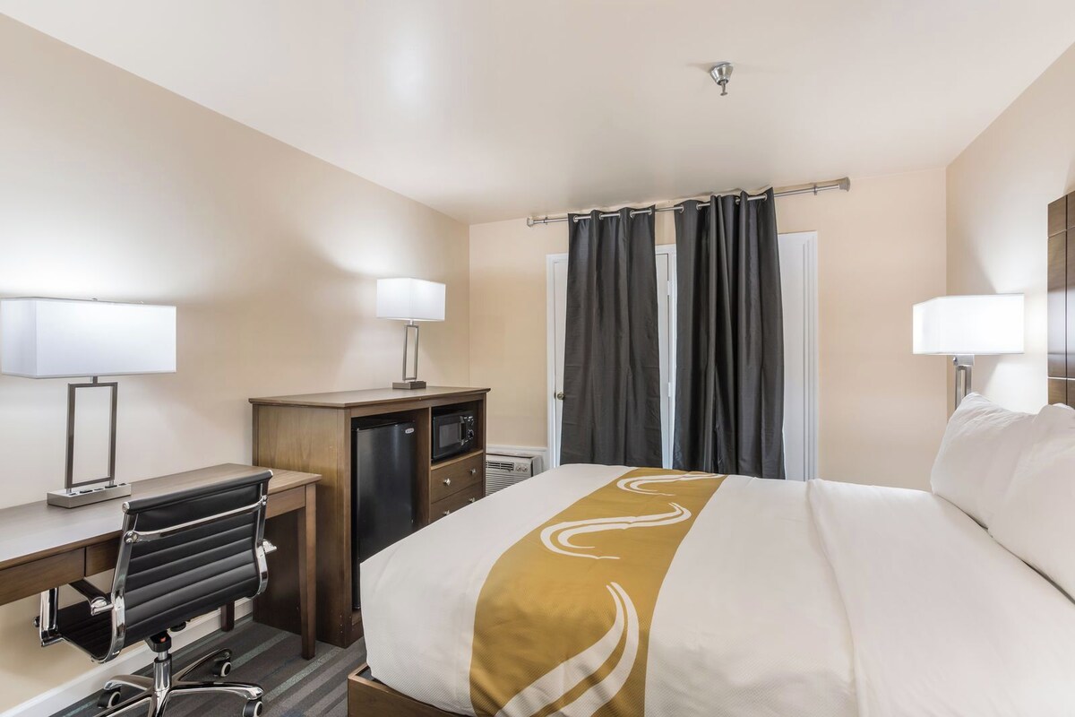 A hotel room with a bed, desk, chair, and a small kitchenette.