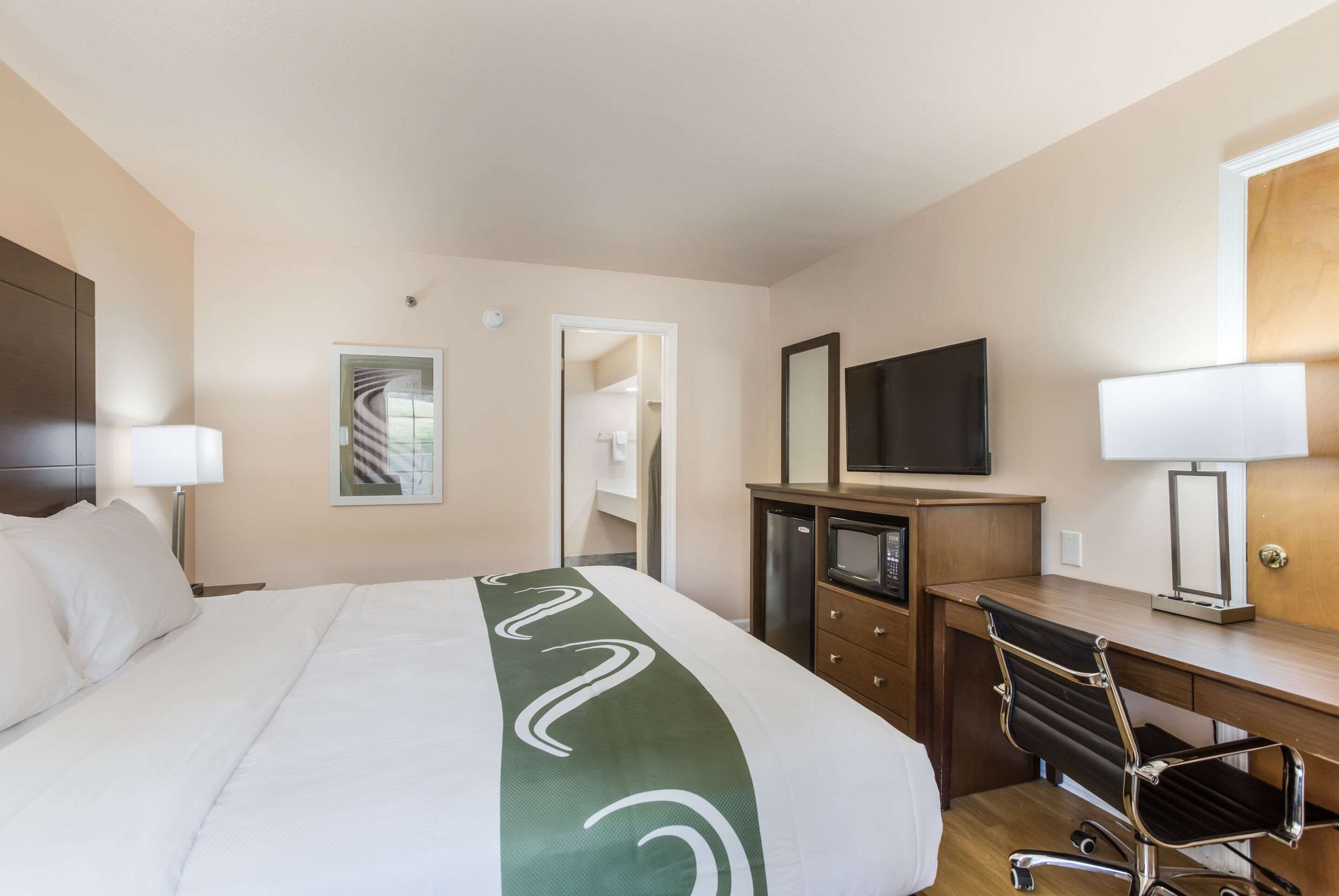 Quarto de hotel com uma cama, escrivaninha, cadeira, televisão e espelho.