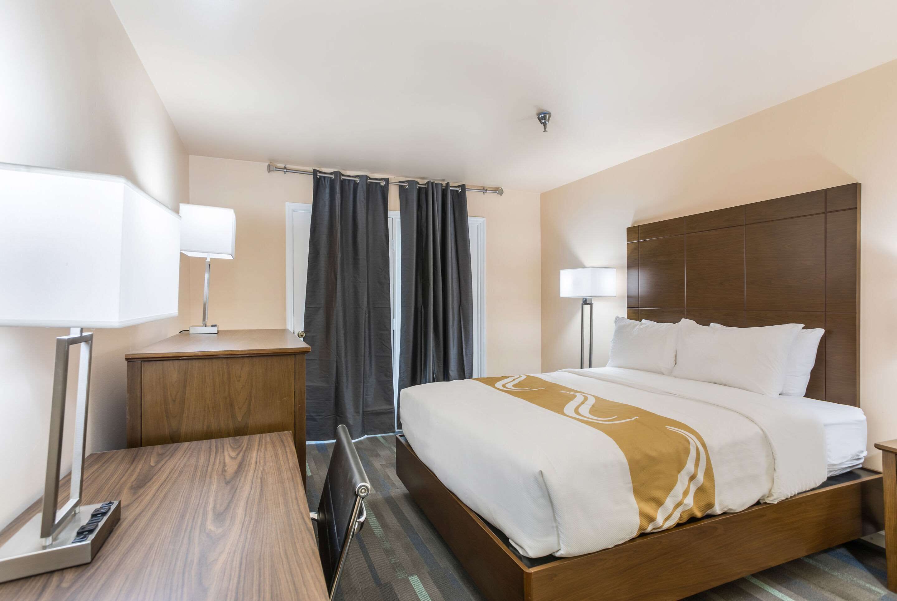 A hotel room with a large bed, a desk, a chair, a lamp, and a curtain.