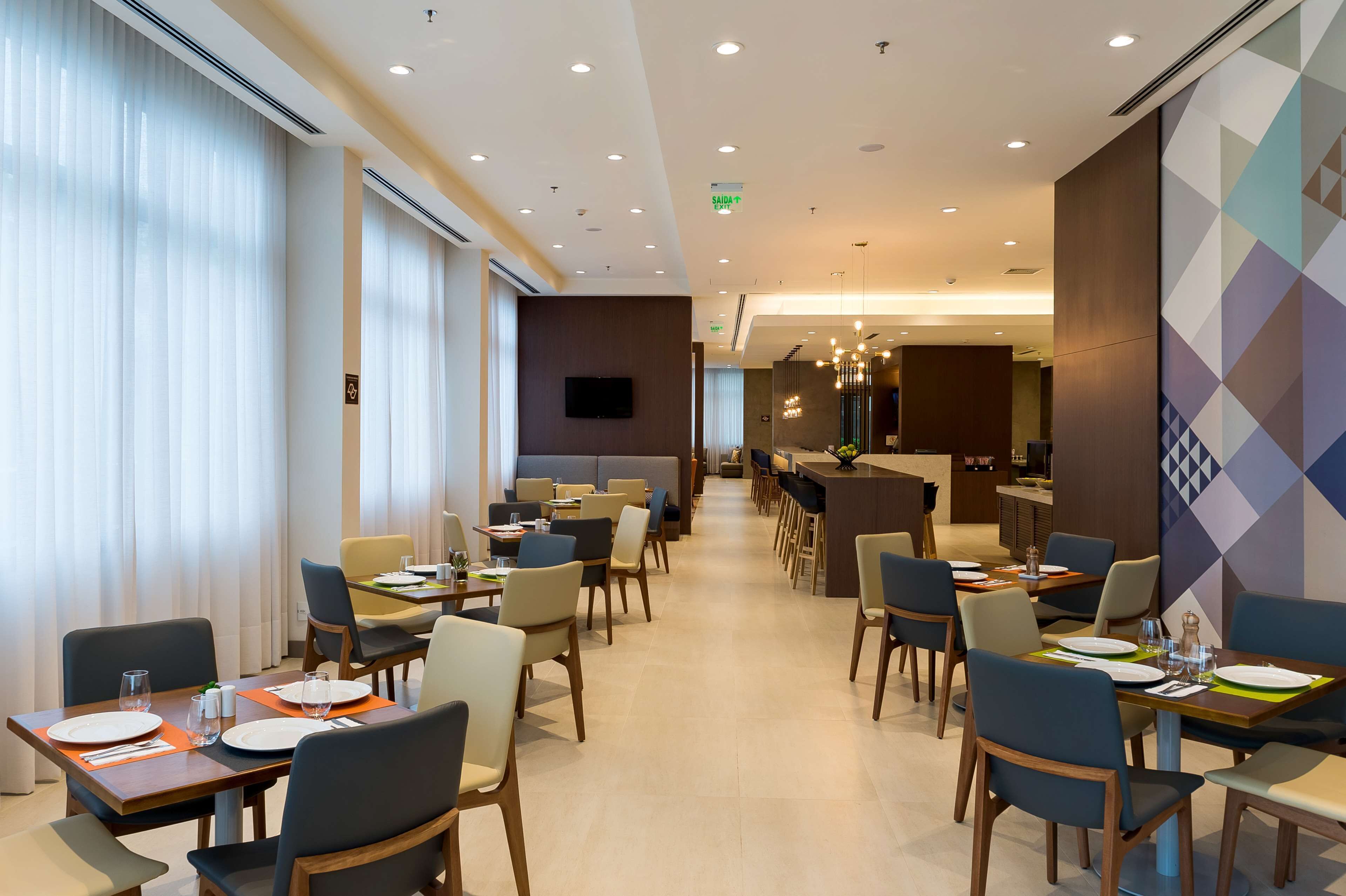A modern hotel dining area with multiple tables and chairs, a flat-screen TV mounted on the wall, and a geometric patterned wall.