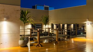 A rooftop lounge with seating and tables, potted plants, and a view of the cityscape at dusk.