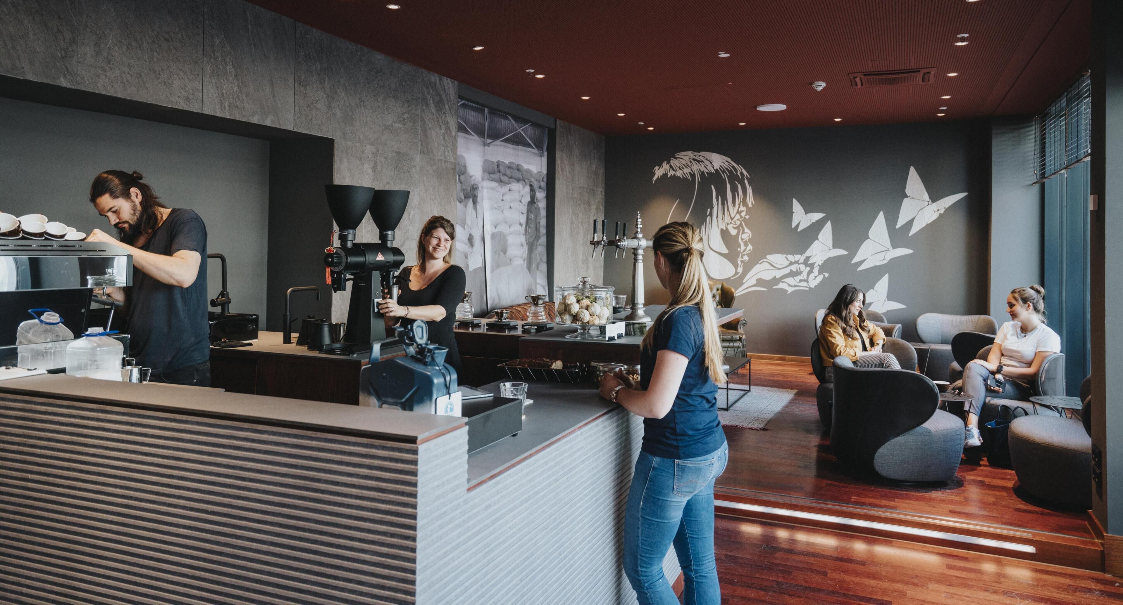 Interior de una cafetería moderna con un barista preparando bebidas, zona de estar y decoración mural.