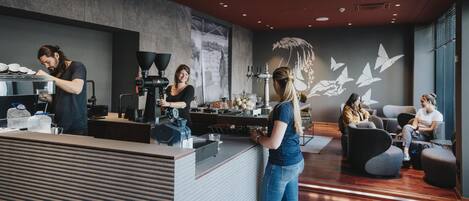 Interior de una cafetería moderna con un barista preparando bebidas, zona de estar y decoración mural.