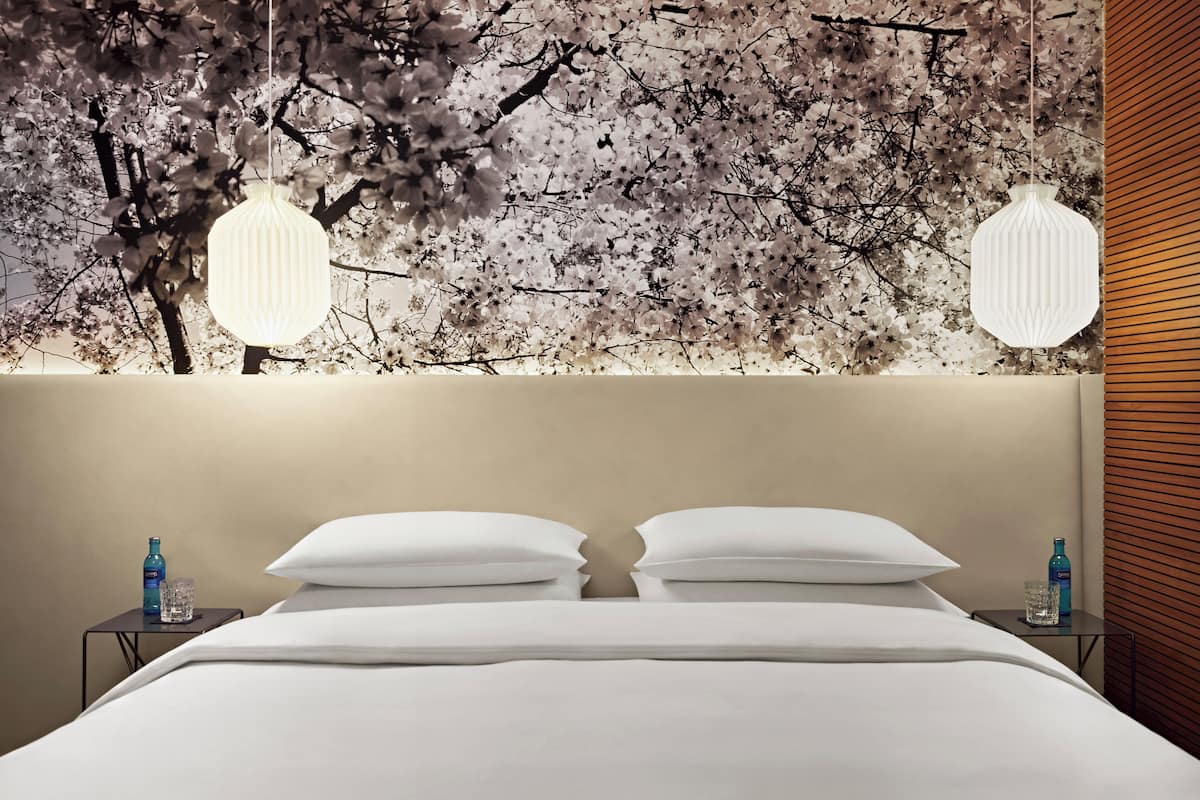 A neatly made bed with white linens and pillows, a bedside table with a water bottle and glass, and a wall with a cherry blossom mural and hanging lights.