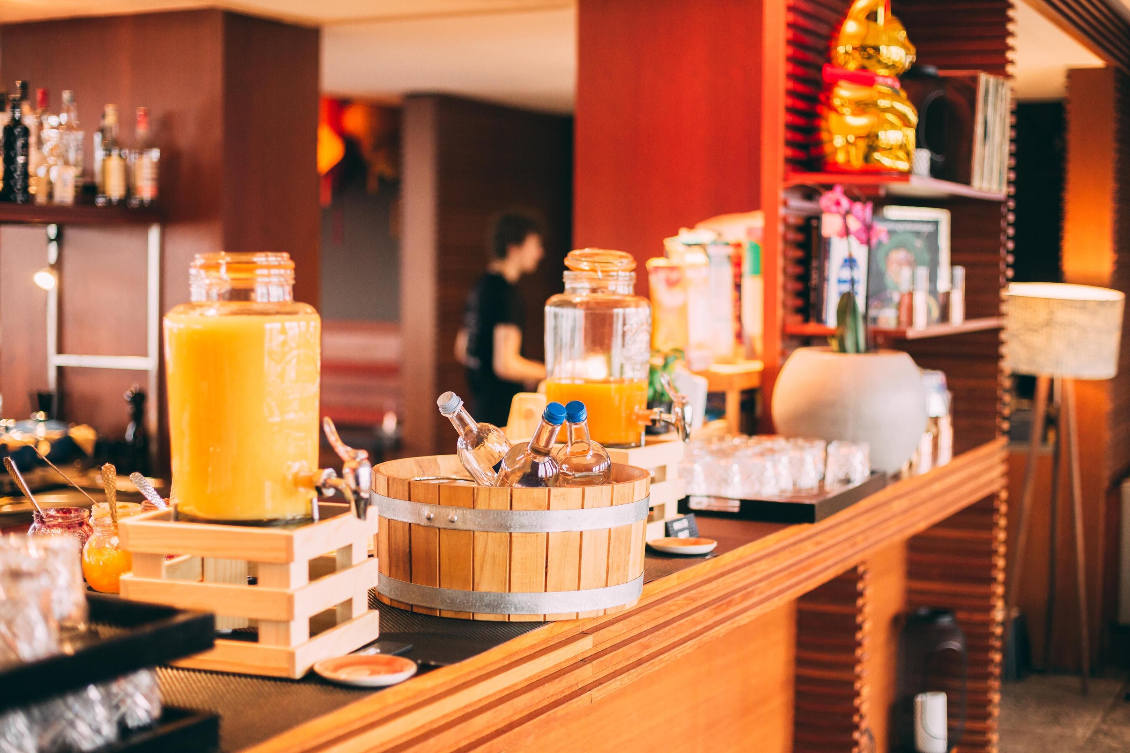 Zona de bar con mostrador de madera, un dispensador grande de bebidas anaranjadas y varias botellas.