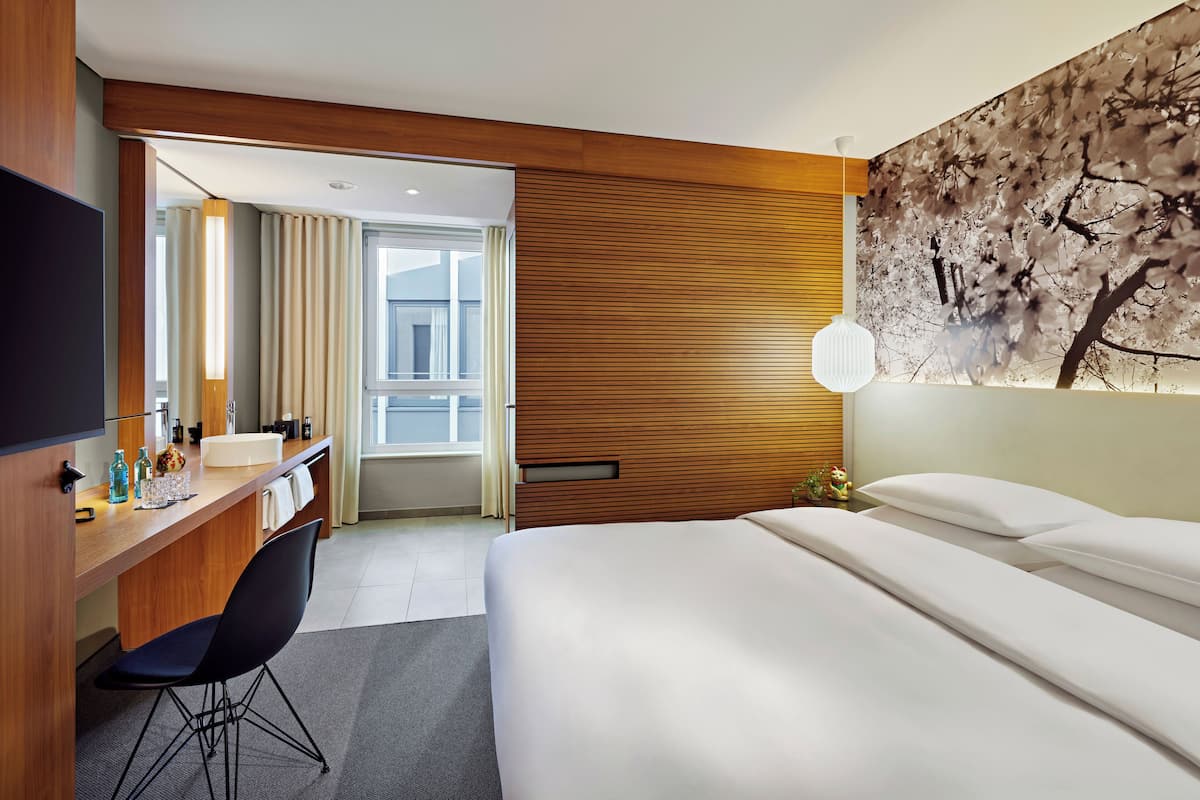 A modern hotel room with a wooden headboard, a large bed, a desk with a chair, a mirror, and a wall-mounted lamp.