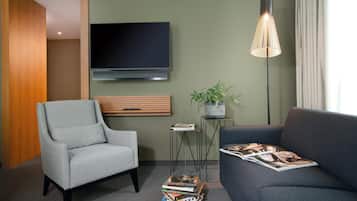 Una sala de estar moderna con un sillón gris, un sofá oscuro, un televisor de pantalla plana, una lámpara de pie, una mesita con una planta y una pila de revistas.