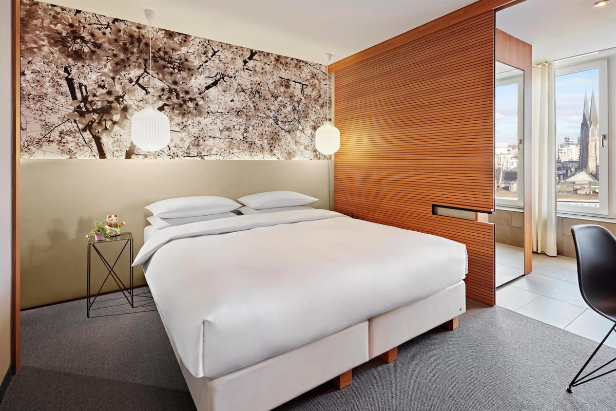 A modern hotel room with a large bed, a wooden panel wall, a city view, and a small side table with a plant.