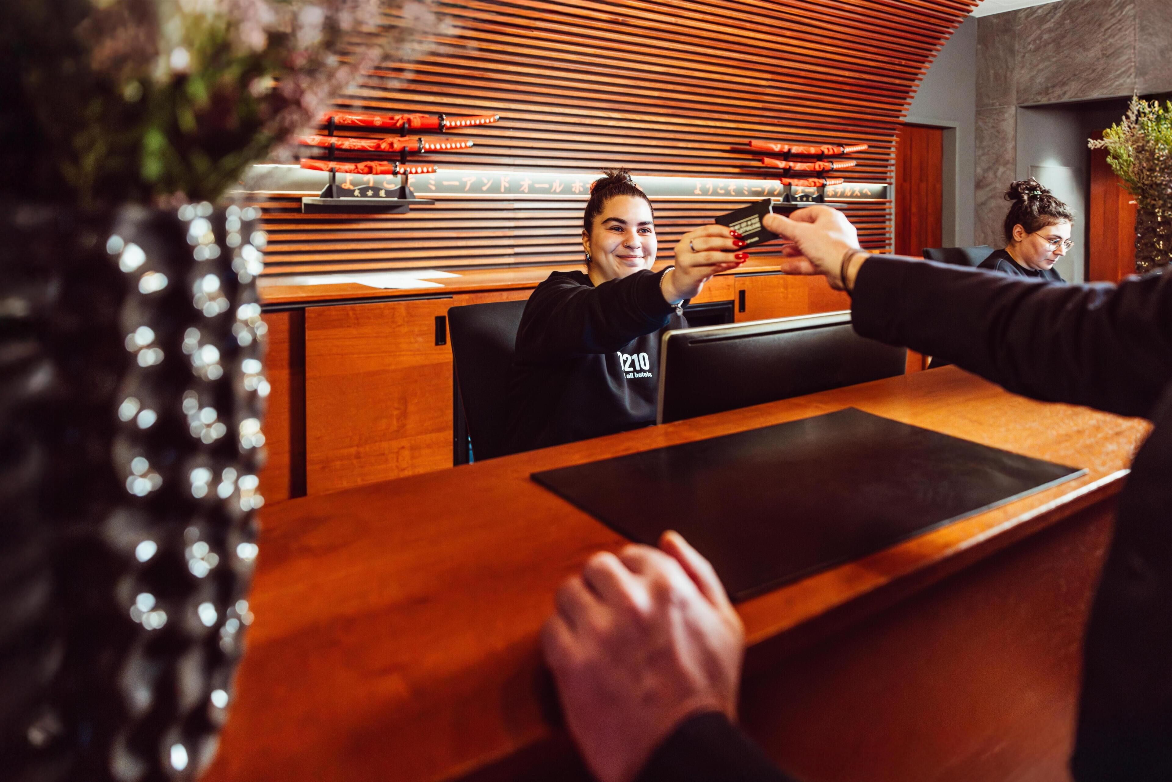 Una reception con scrivania in legno, una sedia nera e una persona che tiene in mano uno smartphone.