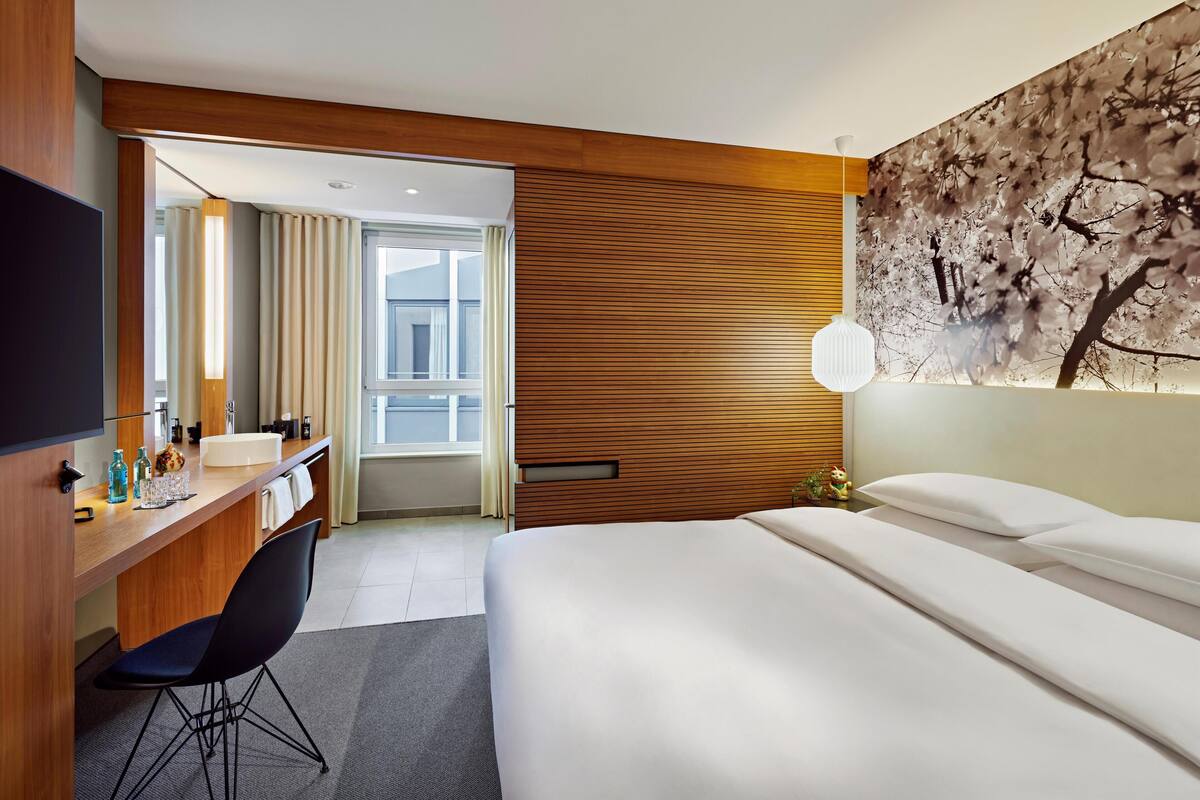 A hotel room with a bed, a desk with a chair, a mirror, a window, and a wall with a floral painting.