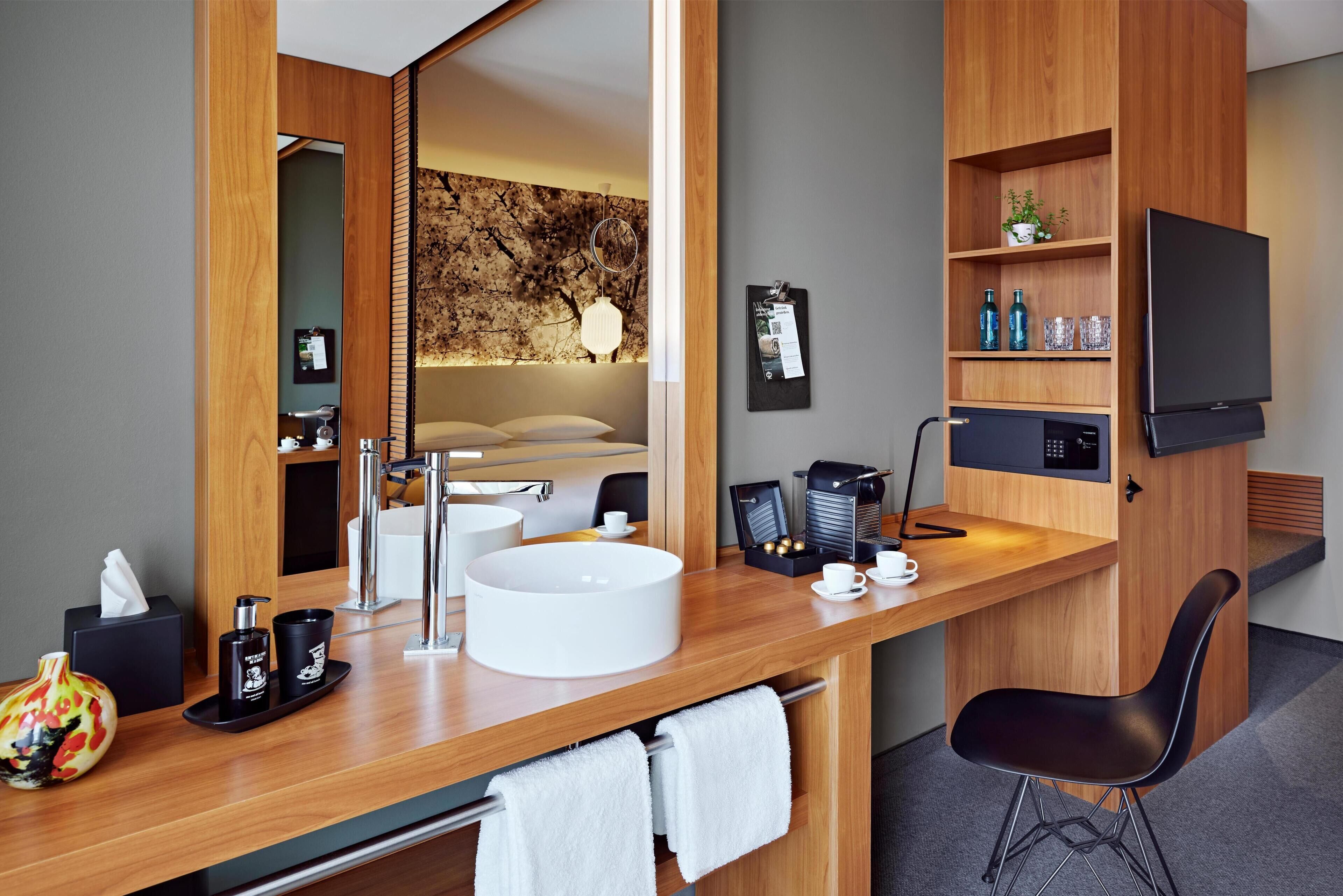 Habitación de hotel moderna con tocador de madera, un espejo grande, un lavabo redondo blanco, una silla negra, televisor de pantalla plana y máquina de café.
