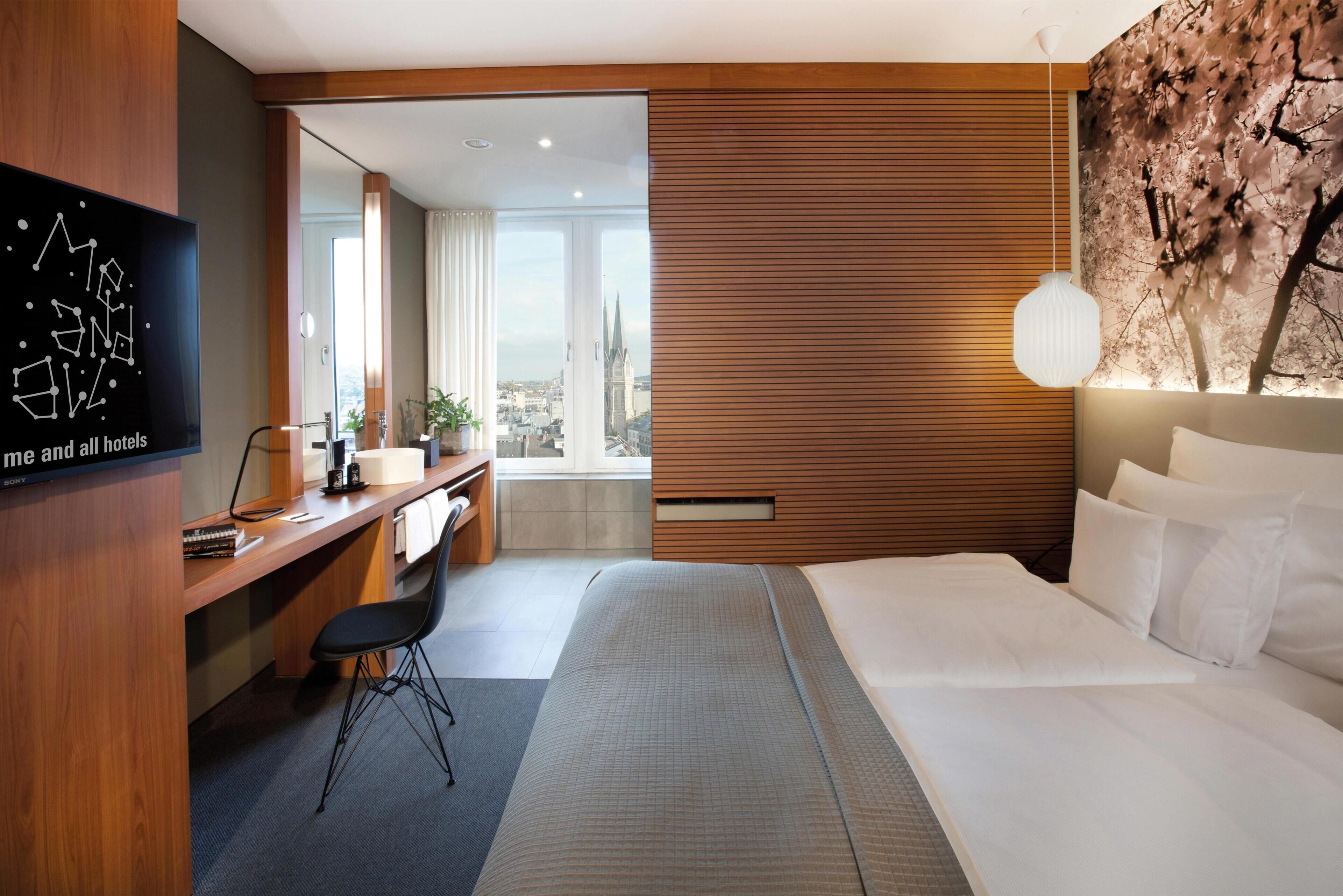 A hotel room with a bed, desk, chair, and a city view through the window.