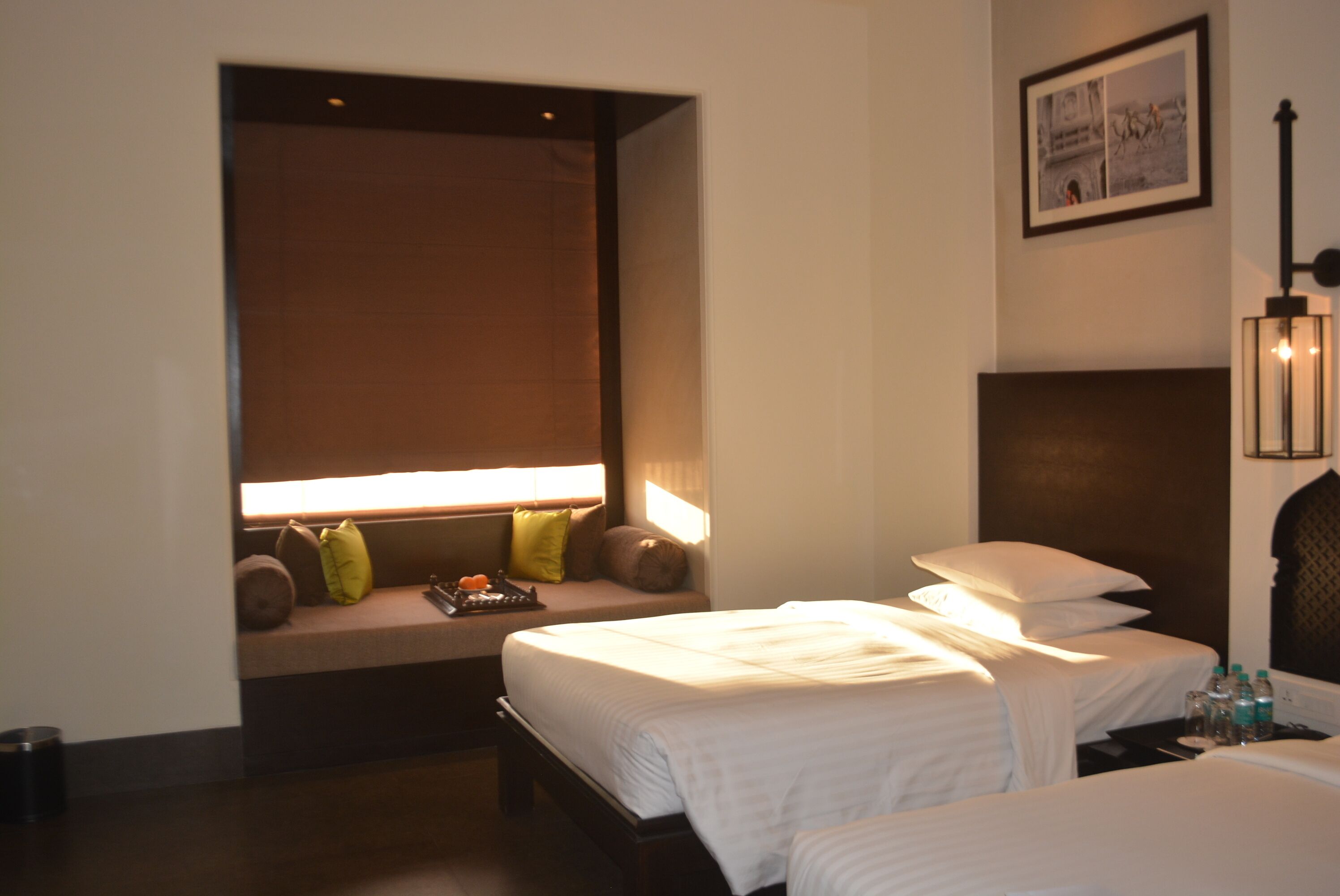A hotel room with two beds, a bench with pillows, a wall-mounted picture, and a bedside table with bottles.
