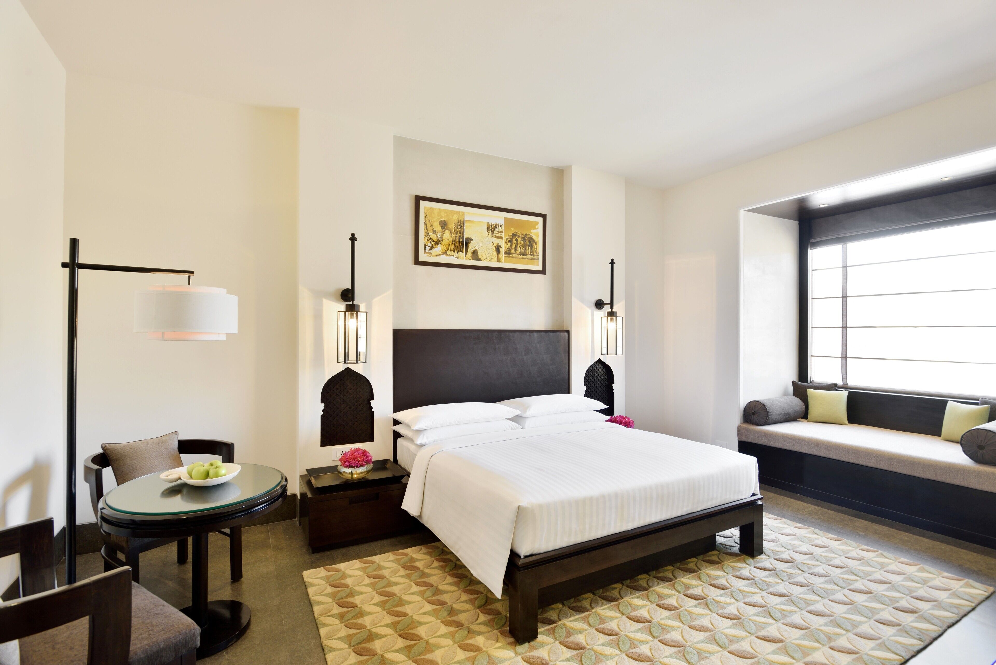 Habitación de hotel con cama, mesita de noche, lámpara, silla, una mesa redonda y una ventana con banco.