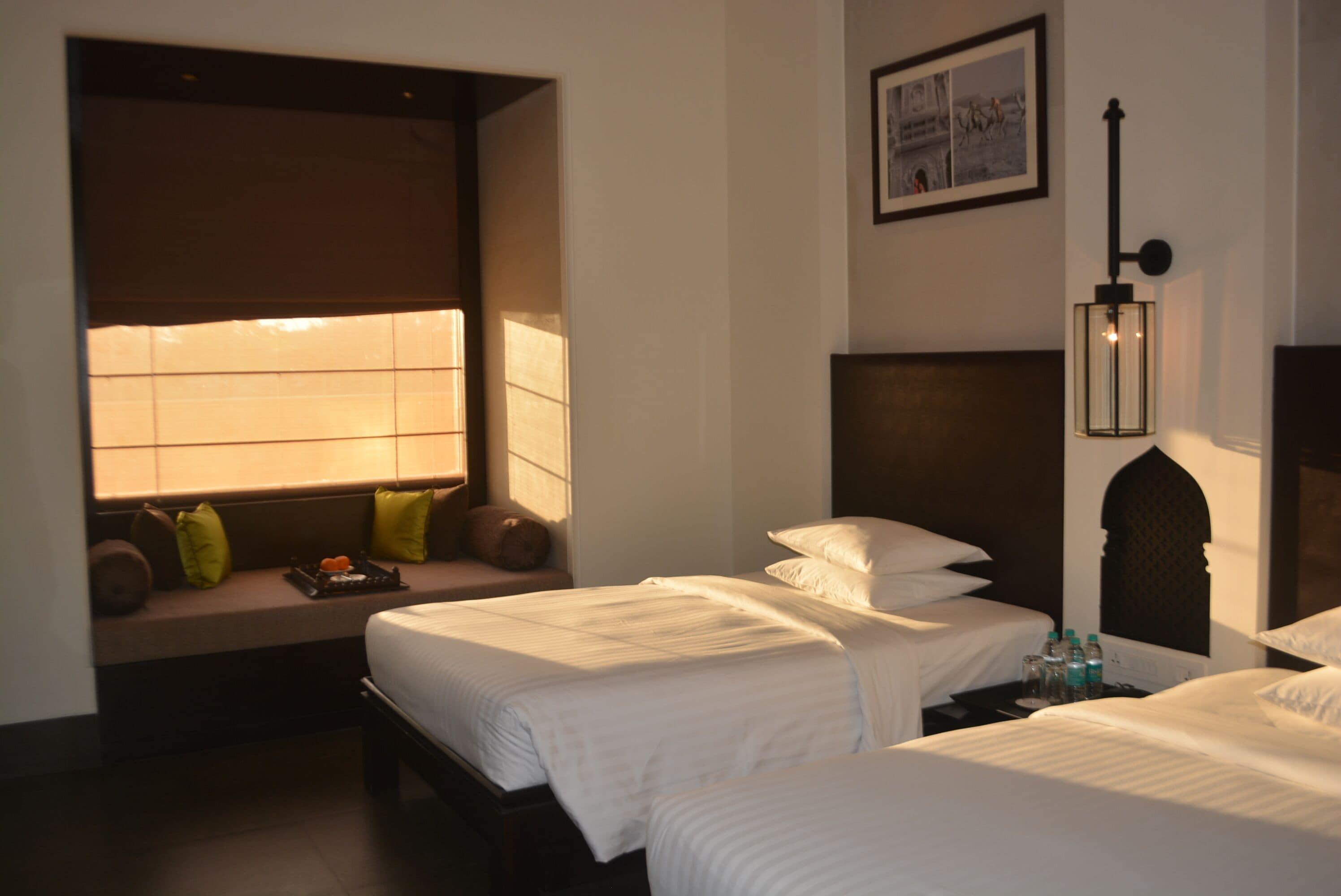 A hotel room with two beds, a window with blinds, a bench with pillows, a wall-mounted light fixture, and a framed picture on the wall.