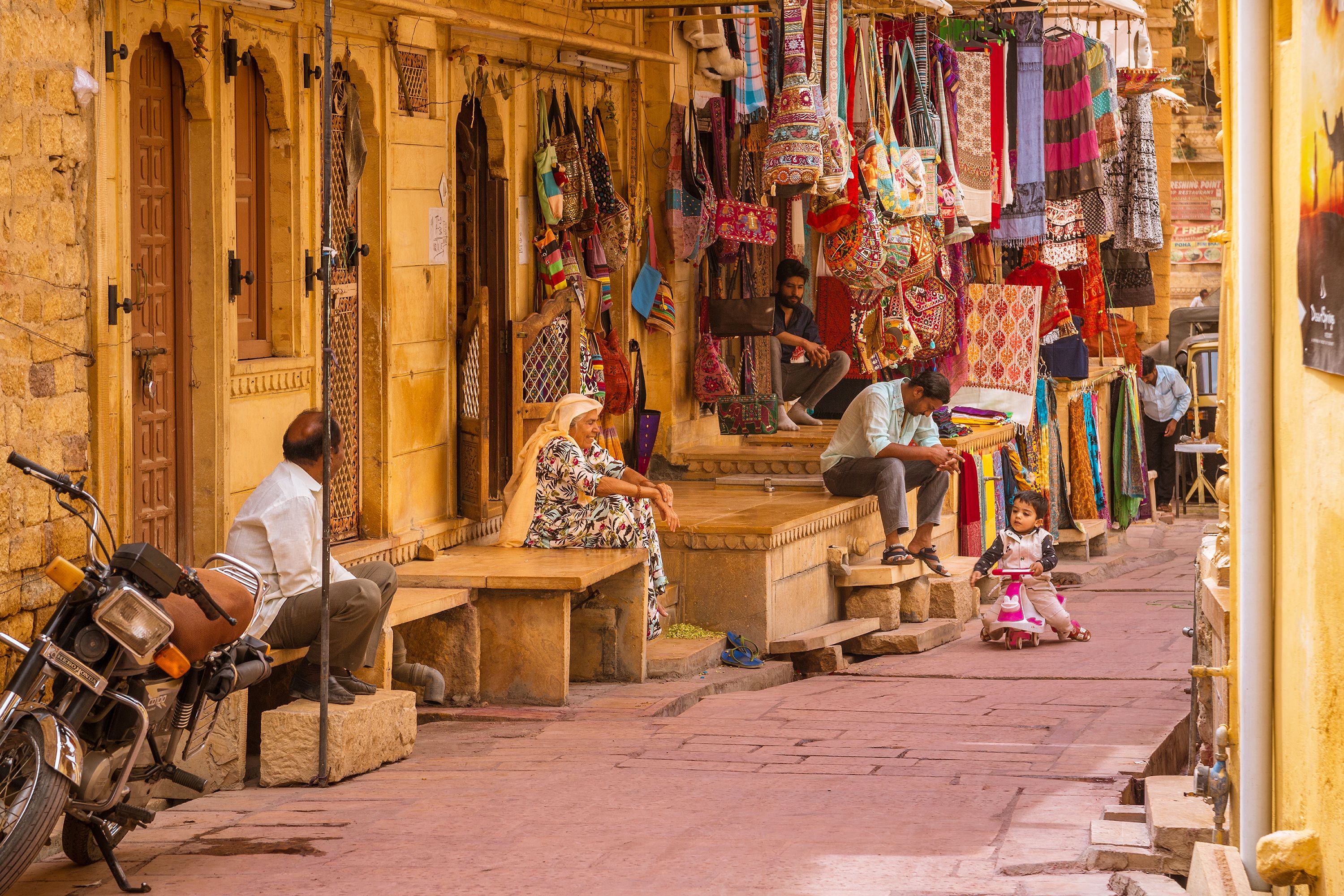 A narrow street with shops on one side and a bench where people are sitting.