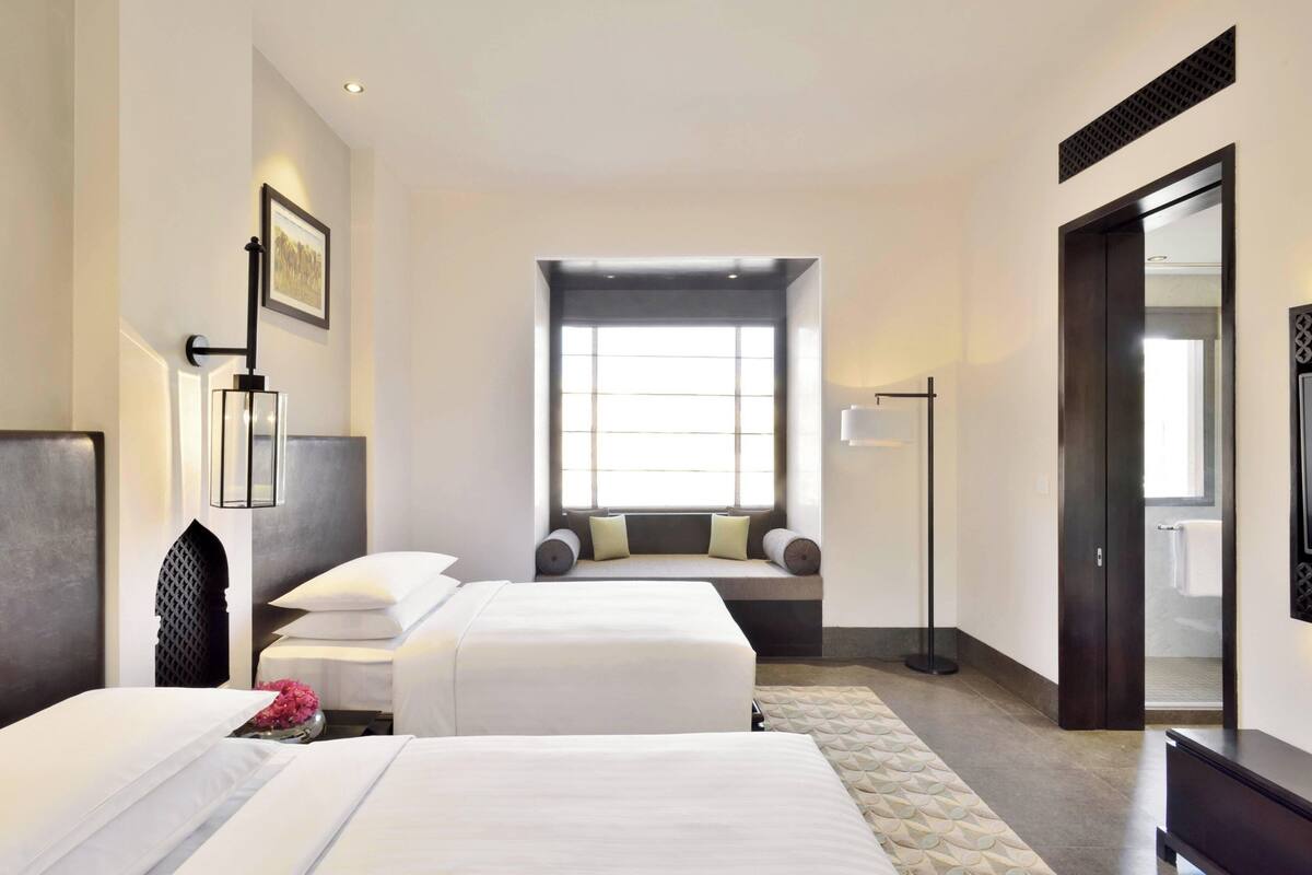 Ein Hotelzimmer mit zwei Betten, einem großen Fenster, einer Sitzecke mit einer Bank und einem Spiegel an der Wand.