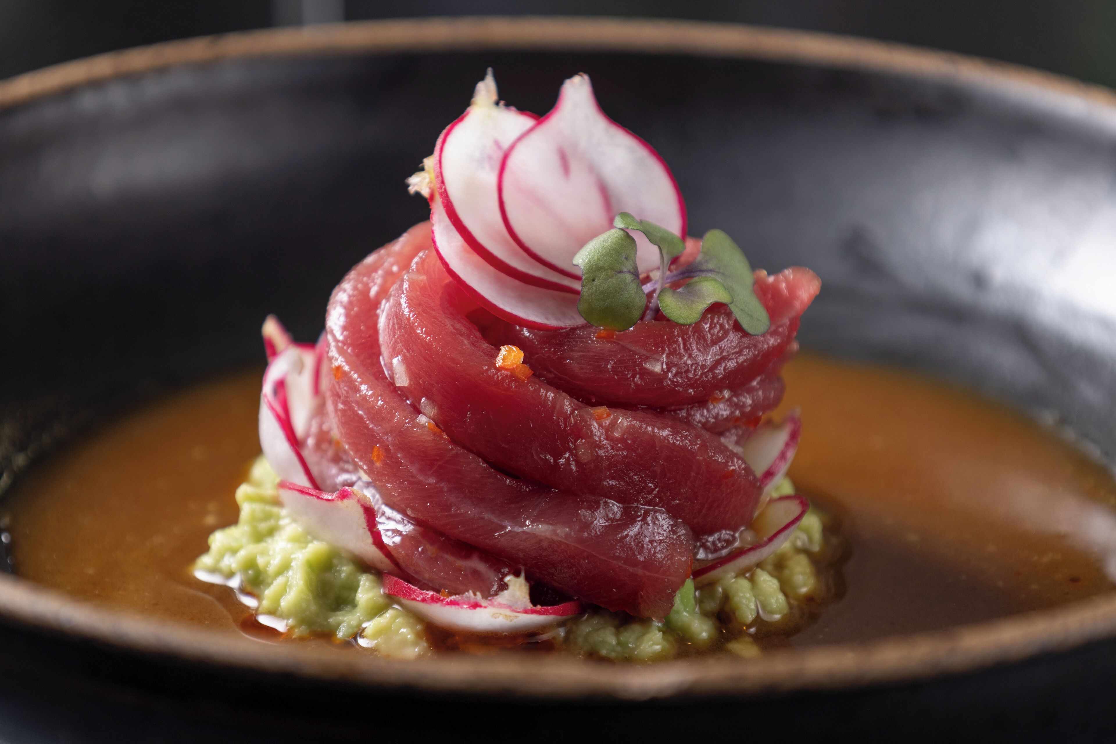 A dish with thinly sliced meat, radish slices, and a green sauce on a dark plate.